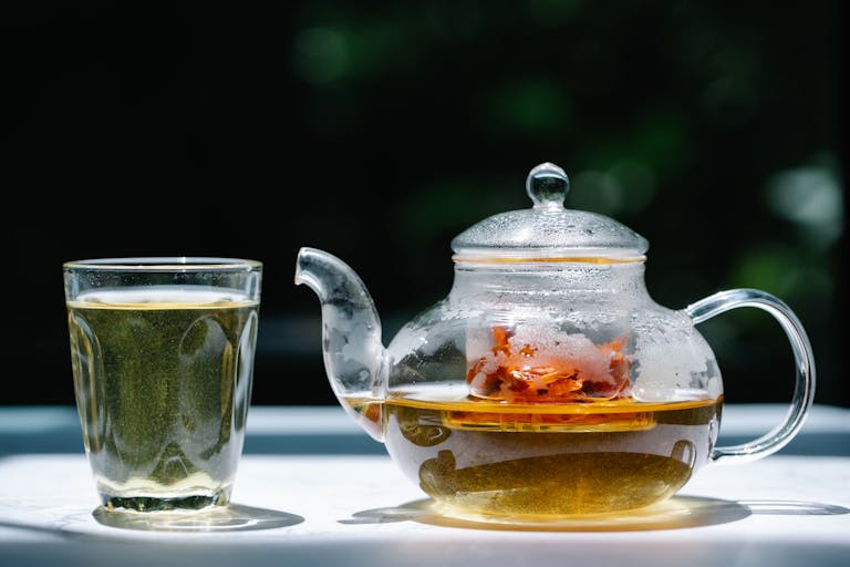 A glass teapot filled with herbal tea next to a transparent glass under sunlight. Refreshing and aromatic.